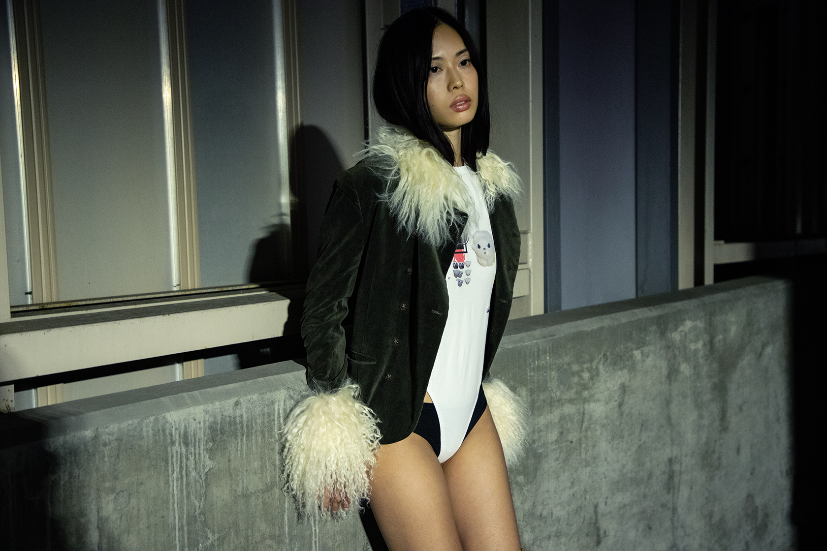 Knee up portrait of a model in a cement parking garage wearing a fur lined black jacket and leaning against a barrier.